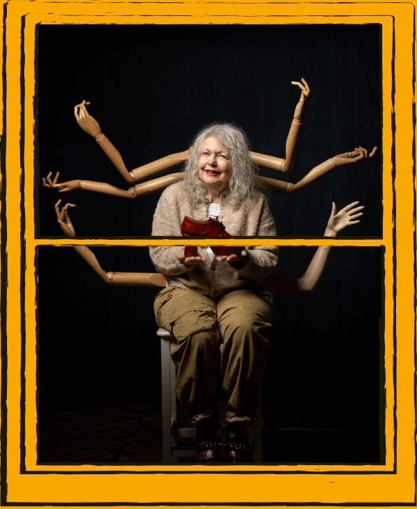 Woman sitting on stool, with mannequin arms behind her, on a black background