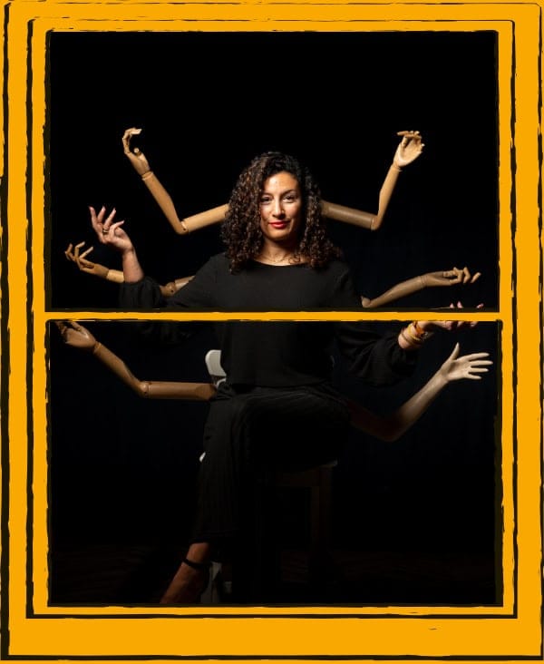 Woman sitting on stool, with mannequin arms behind her, on a black background