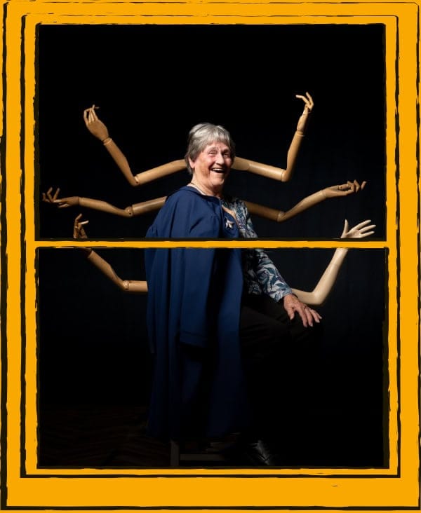 Woman sitting on stool, with mannequin arms behind her, on a black background